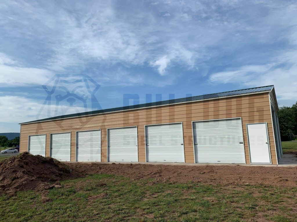Exterior of metal building with six large garage doors, in grassy area with blue sky and white clouds above.